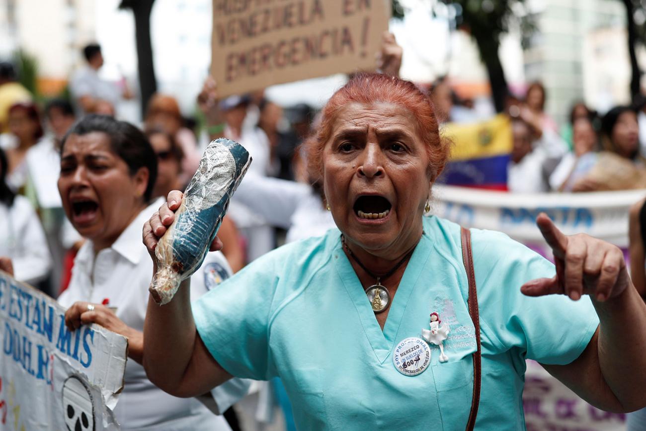 Health care workers protesting shortage of medical supplies outside a hospital in Caracas, Venezuela on November 19, 2019. The picture shows an older woman in the foreground wearing blue hospital scrubs shouting and looking slightly off camera. She is surrounded by other protestors, many of whom are waving protest signs and also shouting. REUTERS/Carlos Garcia Rawlins