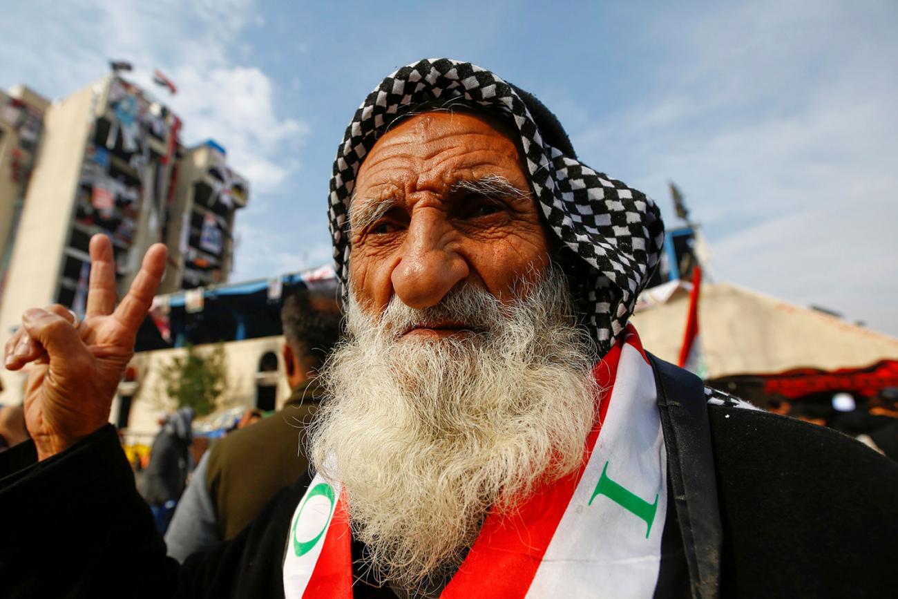 An elderly demonstrator poses for the camera as he makes a V-sign during ongoing anti-government protests, in Baghdad, Iraq, December 10, 2019. He has a black-and-white checkered scarf tied on his head and a red and white sash with green accents, around his neck. He has a very long white beard and friendly eyes. REUTERS/Alaa al-MarjaniSELECT