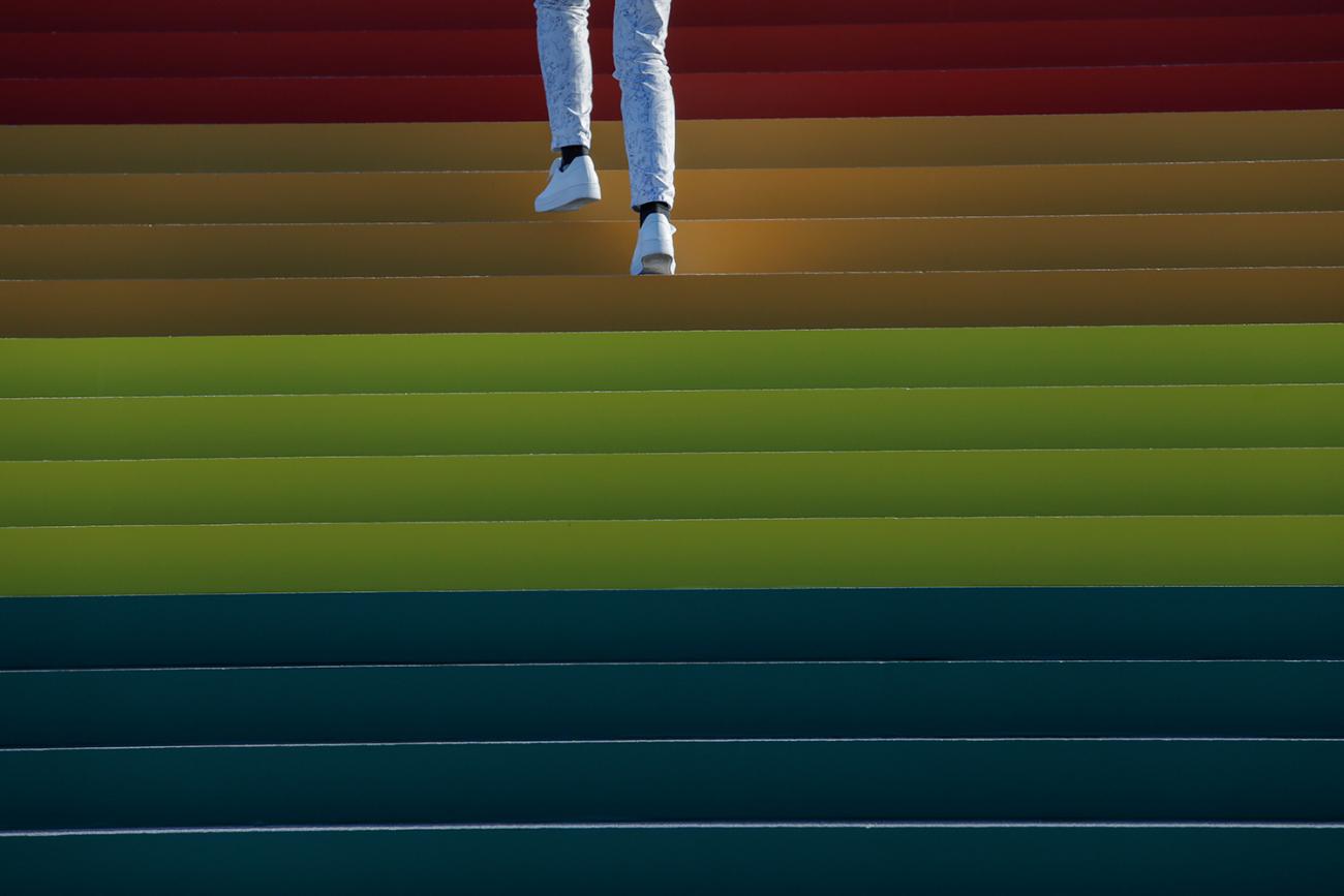 A person walks up a giant LGBTQ+ Pride Flag installed on the steps of Franklin D. Roosevelt Four Freedoms State Park to celebrate World Pride in New York City on June 14, 2019. Photo shows the legs and feet of someone wearing white pants and white sneakers walking up a wide staircase that fills the frame and is brightly adorned in rainbow hues. This is a striking photo. REUTERS/Andrew Kelly