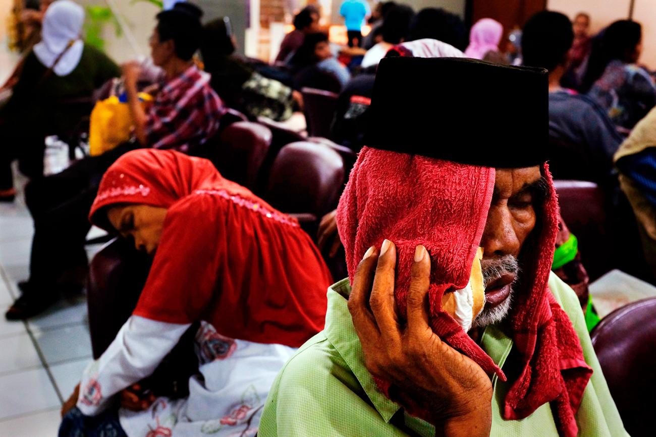 People waiting at Fatmawati hospital in Jakarta on May 17, 2013, after Indonesia announced plans to implement nationwide health care, with the aim to covering all Indonesians by the end of the decade. The picture shows a crowded waiting room with a man in the foreground with a red cloth draped over his head and holding what appears to be a bag of ice against his cheek. He looks like he is suffering. REUTERS/Beawiharta.