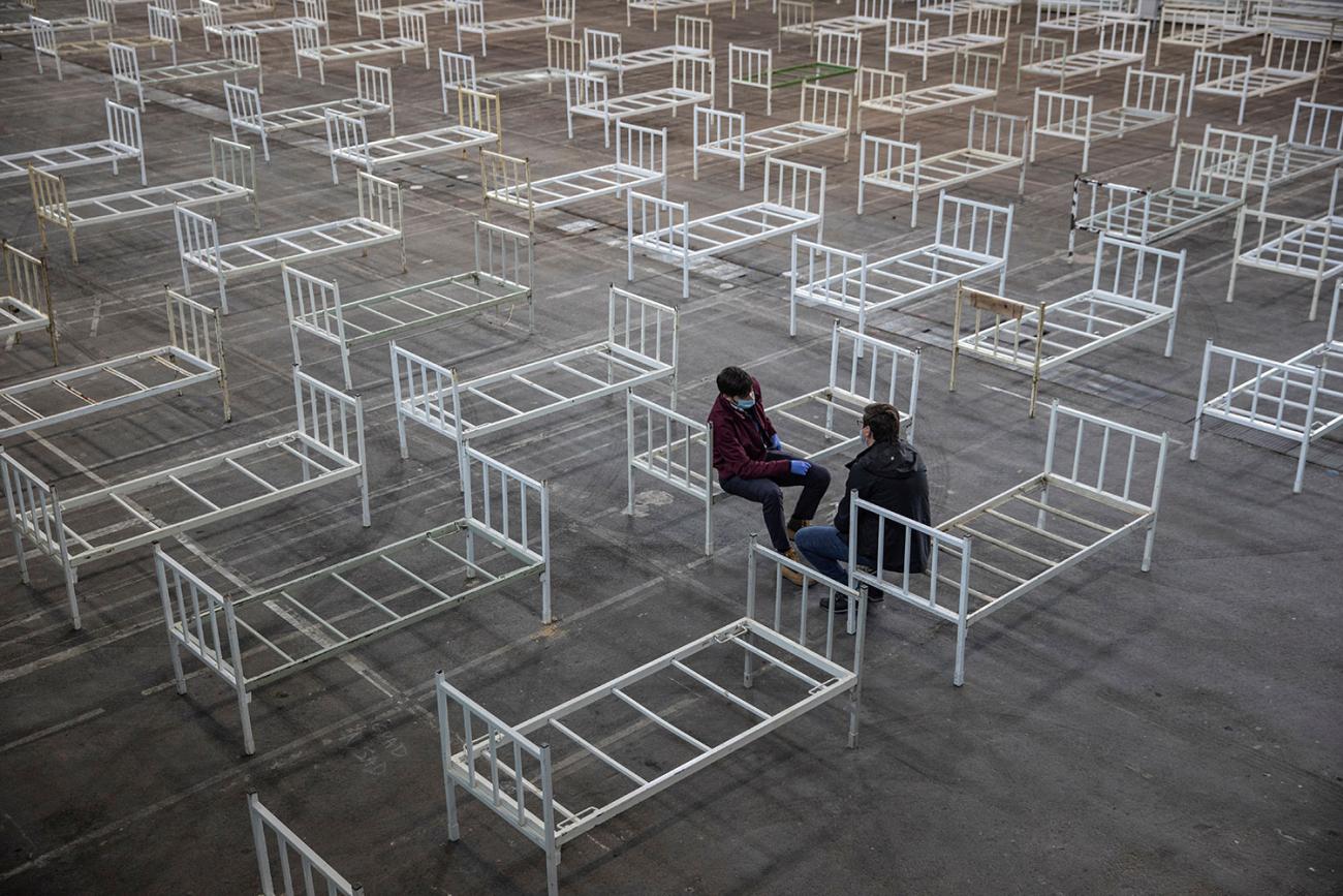 Volunteers take a break as they set up beds inside the Novi Sad Fair to accommodate people who suffer mild symptoms of coronavirus disease (COVID-19) in Novi Sad, Serbia, March 27, 2020. This is a striking image of two people resting on the edge of plain metal bed frames in a large space in which many, many more bed frames are placed. REUTERS/Marko Djurica
