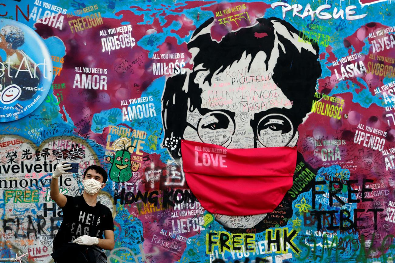 A man wearing a facemask takes a selfie in front of the legendary graffiti-covered John Lennon Wall as the coronavirus pandemic continues in Prague, Czech Republic, April 6, 2020. The photo shows a large graffiti mural of the legendary singer and peace activist with a mask draped over Lennon's face. A mane in the foreground holds his device at arm's length taking a selfie. REUTERS/David W Cerny