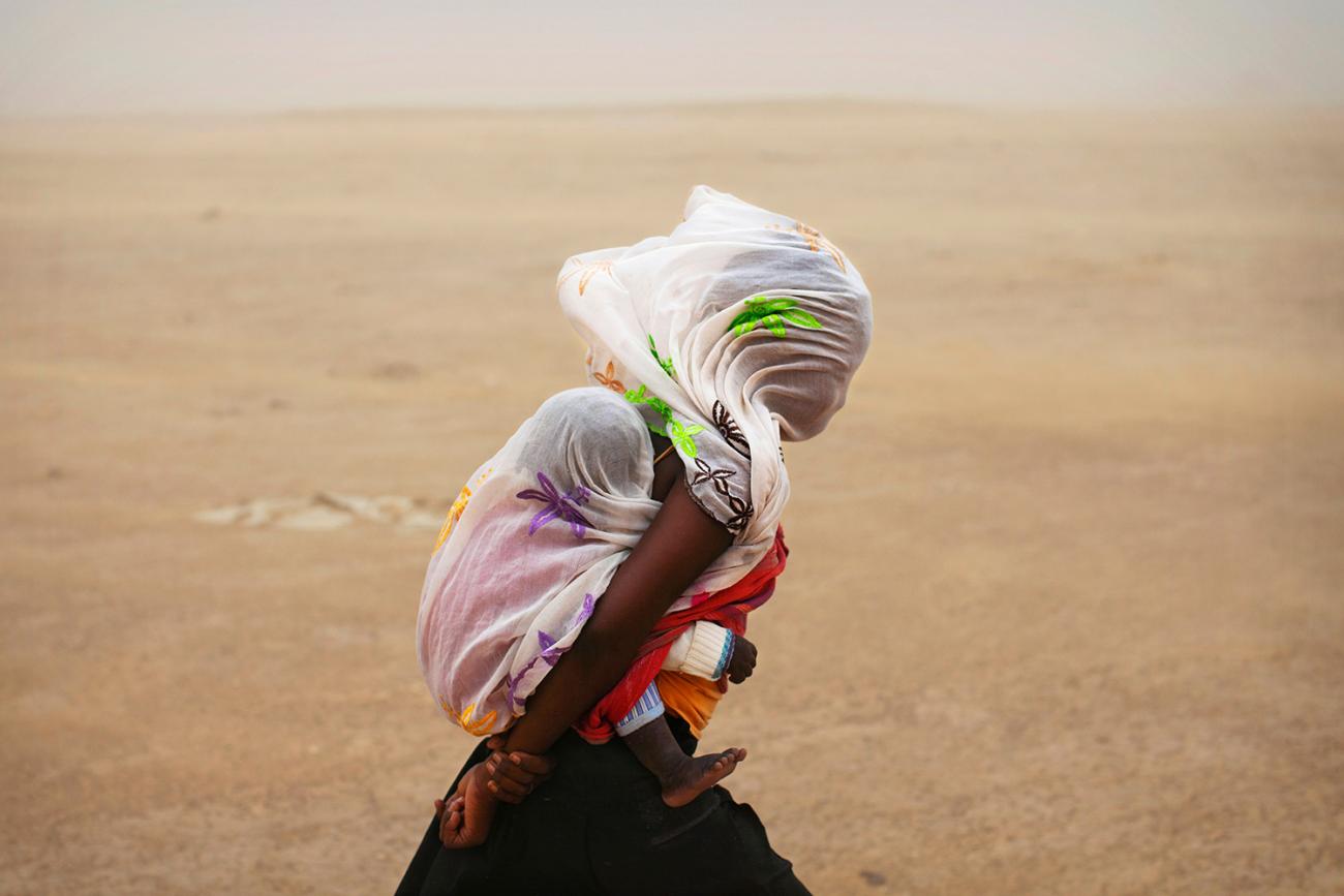 Going out in a storm is a tough choice. Here a woman braves a sandstorm in Timbuktu, Mali, on July 29, 2013. Far worse is the decision many face today whether to brave COVID-19 for other health care.  The photo shows a woman with a baby on her back walking through a desert. The wind is blowing strongly, as evidenced by the billowing cloth on their heads. REUTERS/Joe Penney