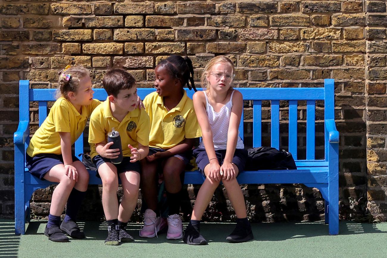 Four of the world's two billion children under the age of fourteen sit on a bench in the playground at St John's Primary School in in London on July 16, 2020 amid the spread of coronavirus. The photo shows four kids on a bench. One of them is looking at the camera. REUTERS/Kevin Coombs