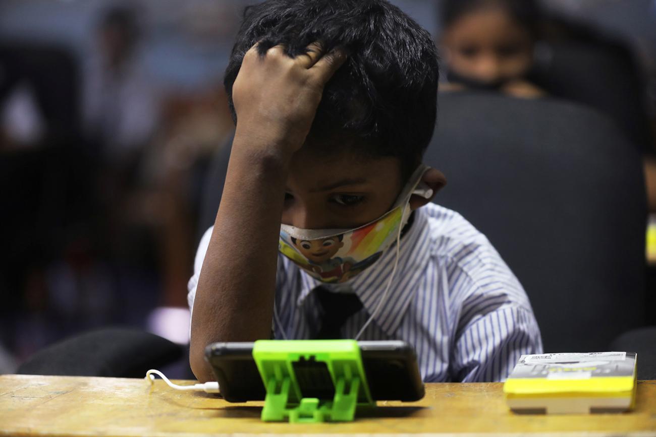 A student watches an online lecture in a digital mobile education library—which provides mobile phones to children who lack access for education during COVID-19 in Mumbai, India, on October 16, 2020. The photo shows a small child at a school desk with a mobile phone on the table in front of them. REUTERS/Francis Mascarenhas
