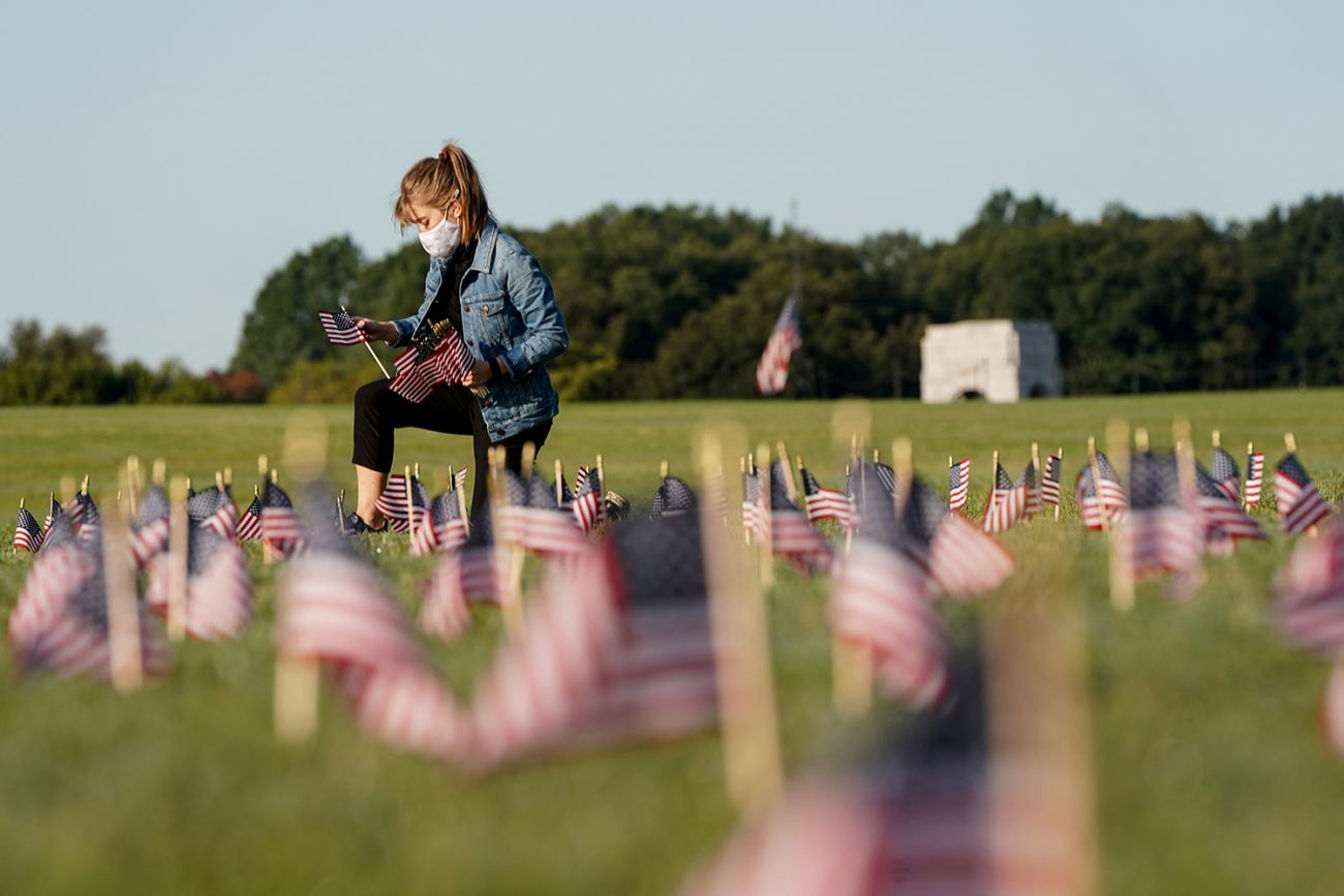 A volunteer places American flags on the National Mall in Washington DC—representing some of the more than 200,000 lives lost in the United States to coronavirus—on September 22, 2020. The photo shows a young woman on one knee planting a small American flag in the ground. Many more can be seen blurred and out of focus in the foreground. REUTERS/Joshua Roberts