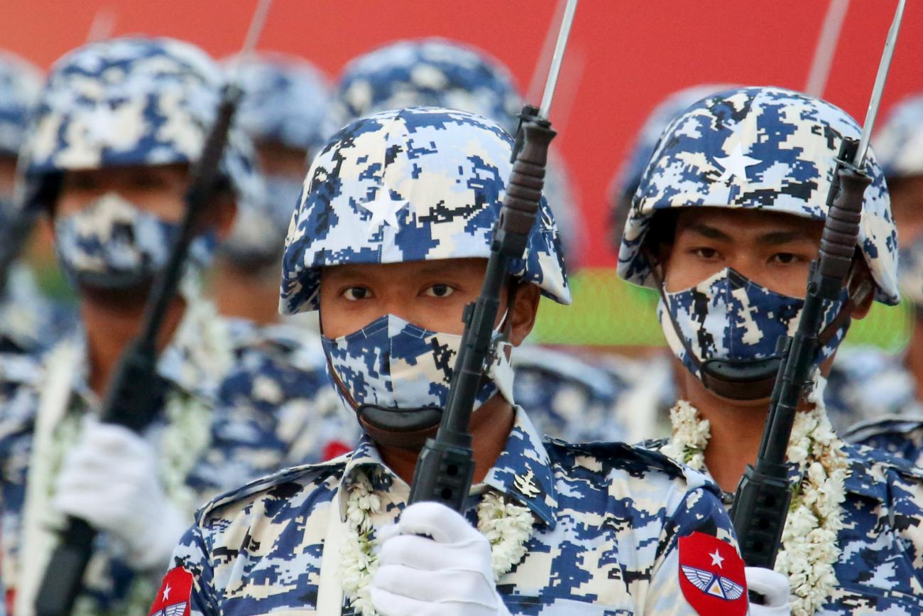 Soldiers in blue camouflage helmets and face masks stand in formation holding bayonets.