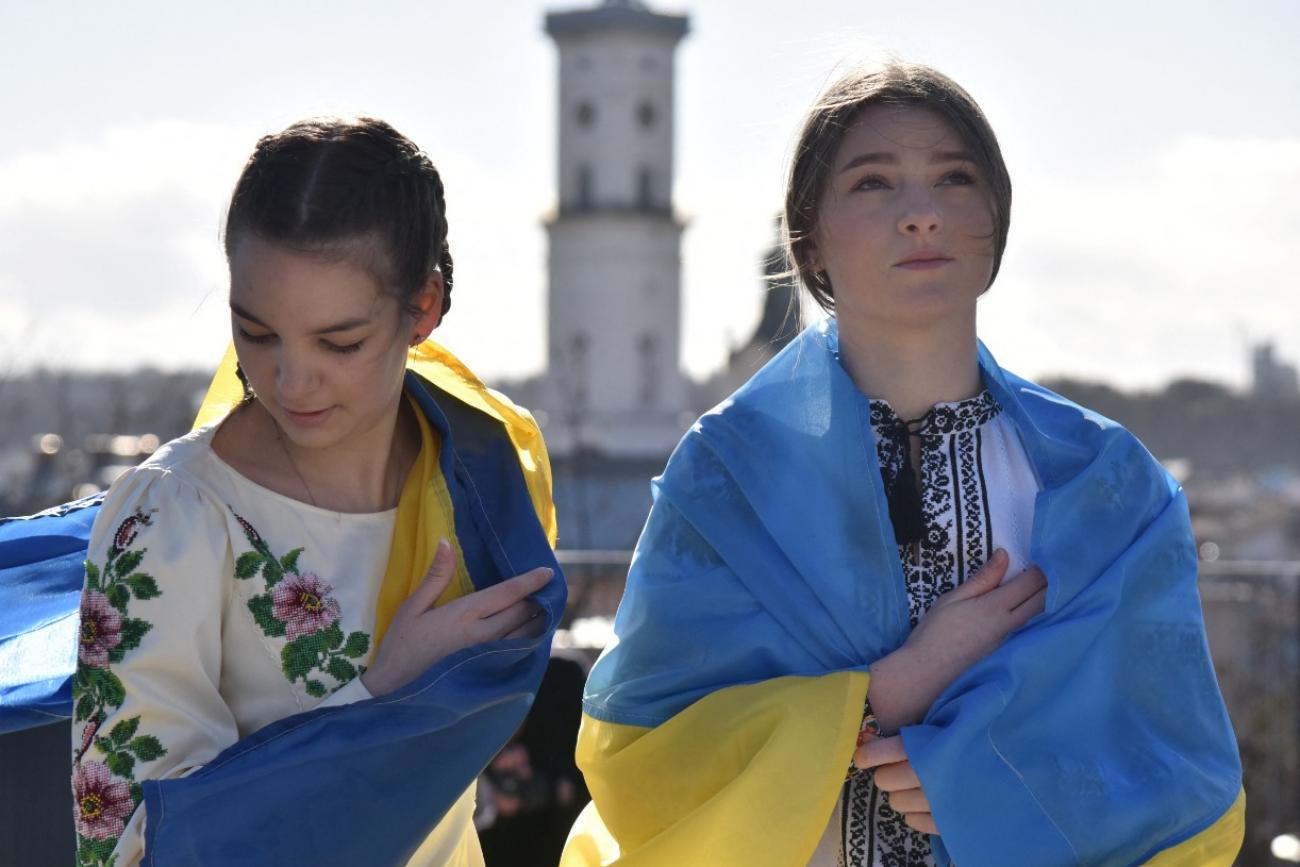 Two girls stand in Lviv, Ukraine wearing traditional Ukrainian blouses and wrapped in the blue and yellow Ukrainian flag