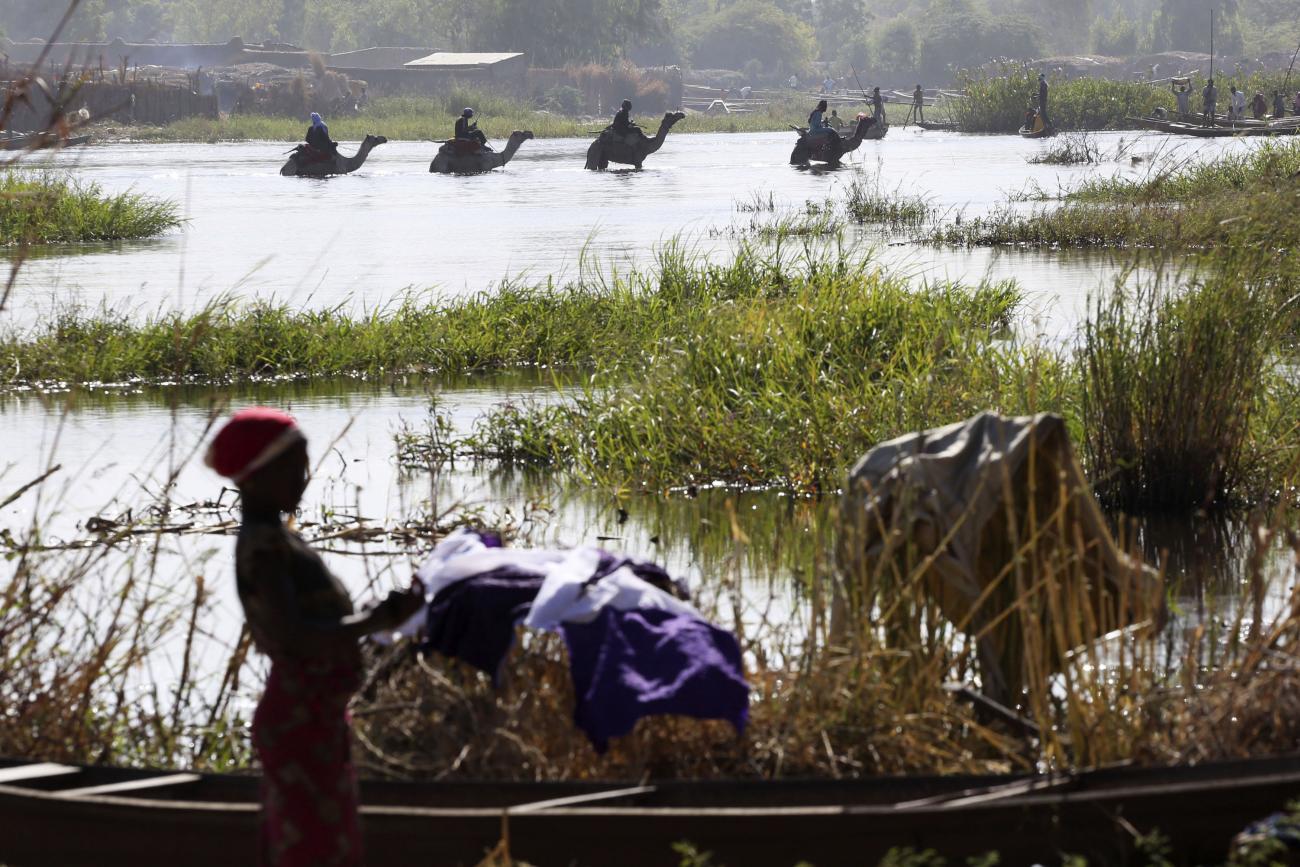 A woman does laundry in Lake Chad. In the background, men on camels wade through the water. 