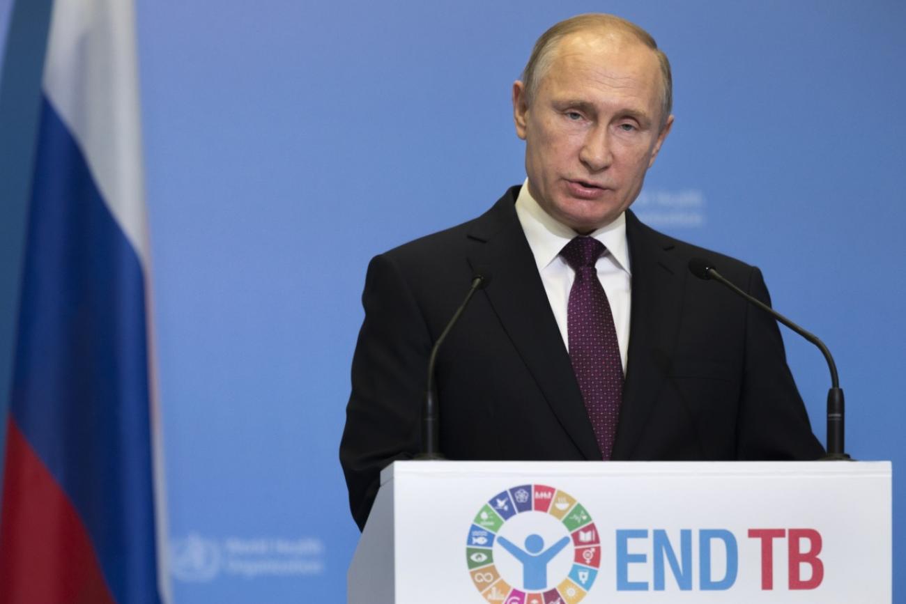Russian president Vladimir Putin stands in front of a podium that says "End TB" with a bored expression on his face.  