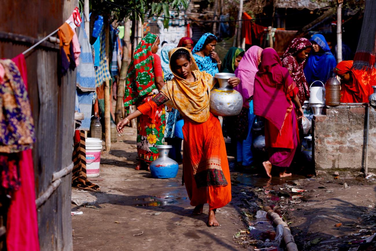 A woman in an orange dress carries a water jar after collecting water from a common water tap. In the background are other women in colorful garments who don't have direct access to water at home in Dhaka, Bangladesh, February 21, 2022. 