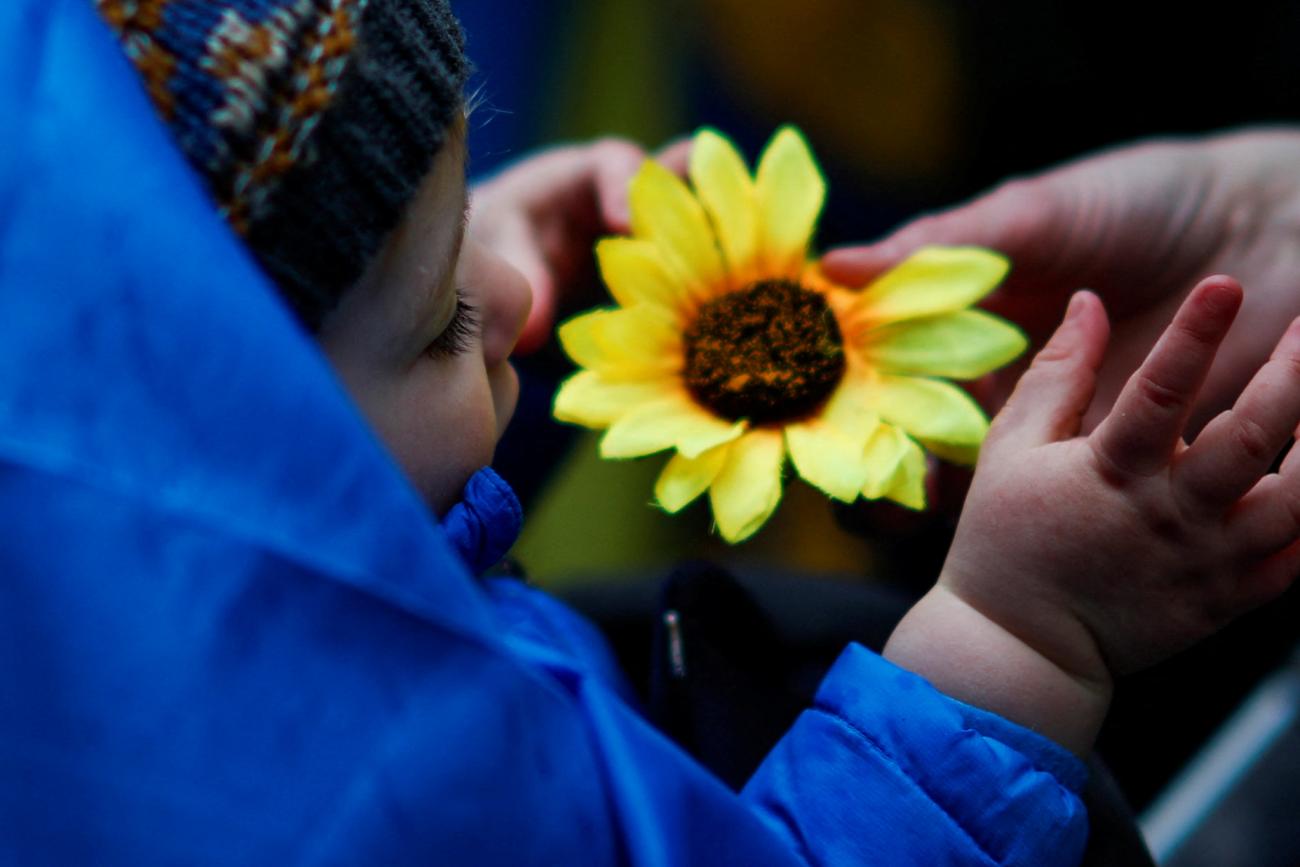 A baby in a blue coat plays with an artificial yellow sunflower. The colors evoke the blue and yellow of the Ukrainian flag. 