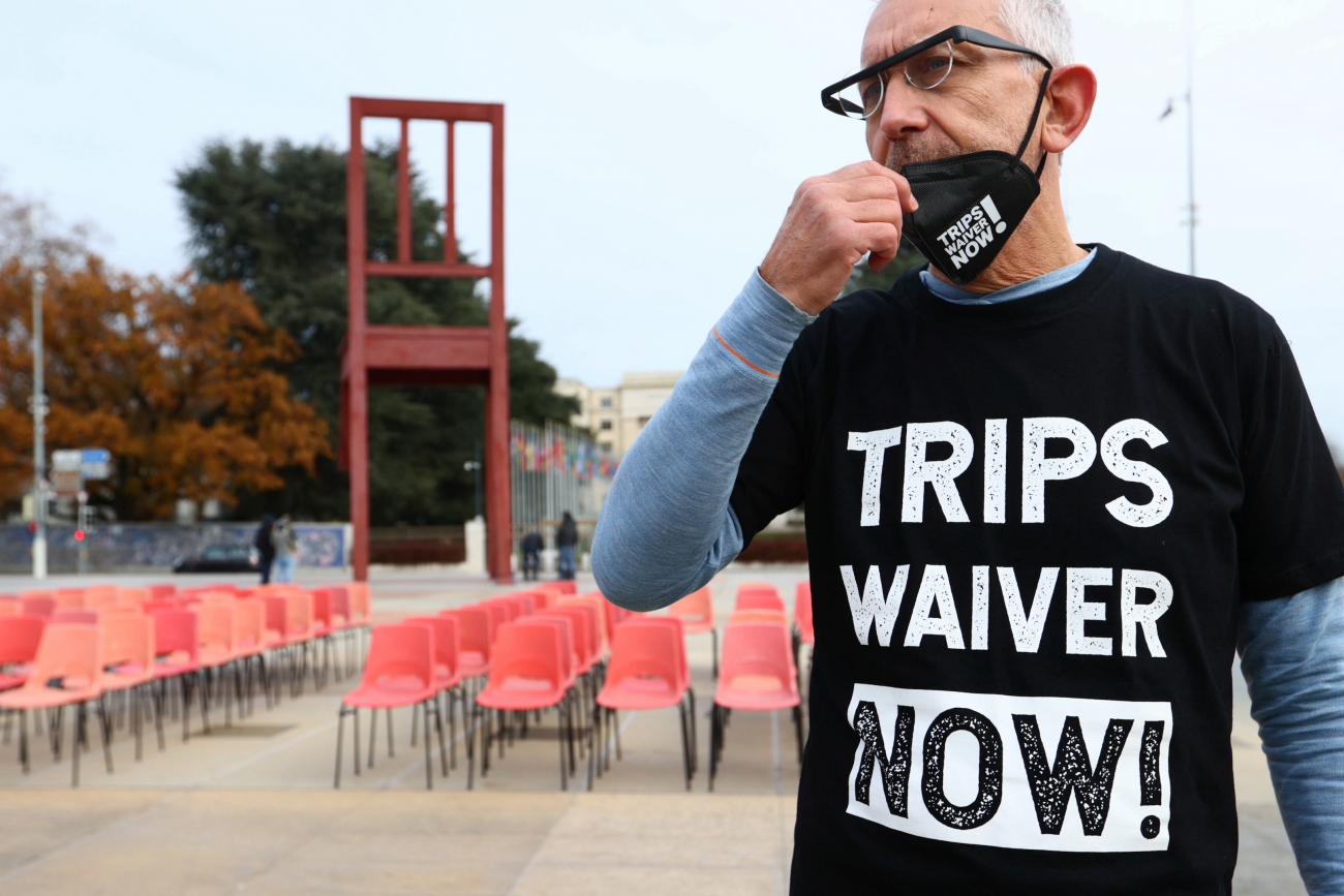 A man adjusts a face mask and is wearing a black shirt that reads "TRIPS WAIVER NOW!" He stands in front of rows of empty chairs.