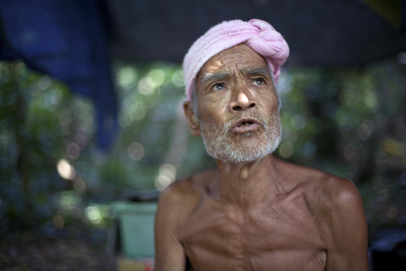 A shirtless elderly man with tanned skin and a white beard wearing a lavender turban is captured mid-speech