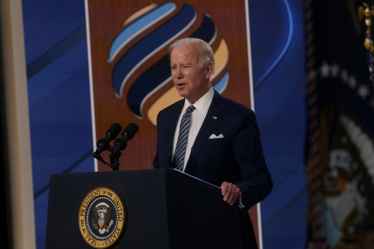 President Joe Biden delivers a speech at a wooden podium while wearing a dark blue suit with a white shirt and striped tie