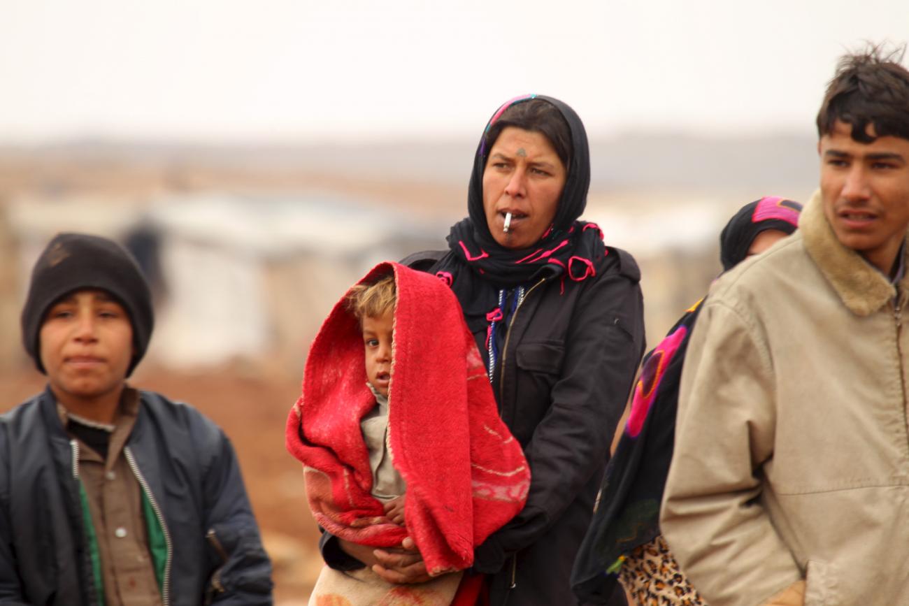 An Syrian woman with a serious expression on her face dressed in dark colors holds a child who is partially obscured by a bright orange scarf draped over his head. On either side of her, are young men in winter coats walking with her through a desert landscape