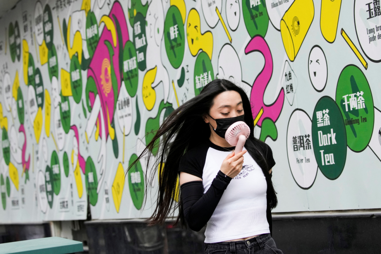 A woman in a white and black t-shirt wears a face mask uses a handheld fan as she walks on a street on a hot day, following the coronavirus disease (COVID-19) outbreak in Shanghai, China July 19, 2022. A colorful white, green, pink and yellow mural is pictured in the background.