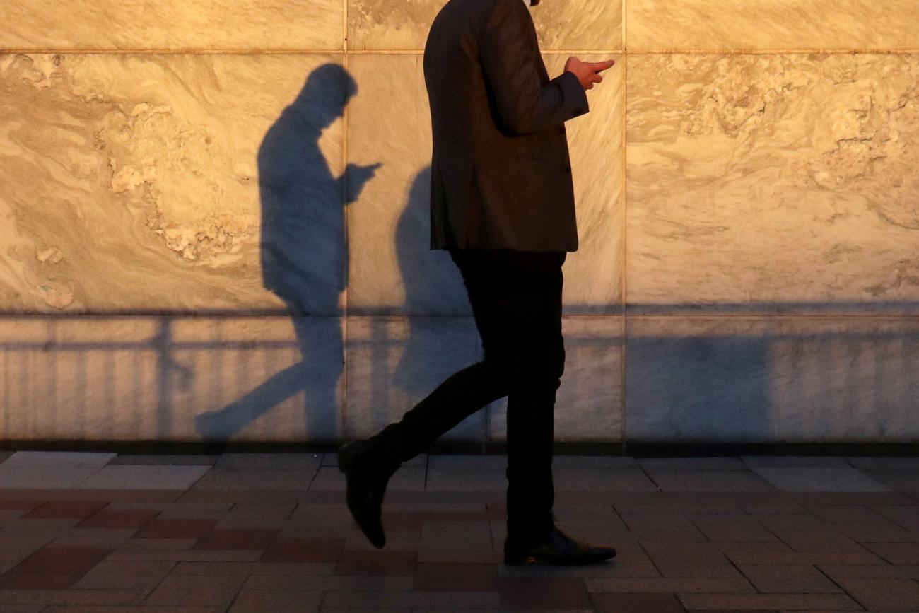 A man using a smart phone walks through London's Canary Wharf financial district in the warm yellow evening light, casting a shadow in London, United Kingdom, on September 28, 2018.