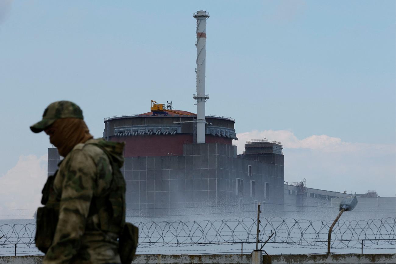 A Russian soldier in a camouflage uniform bearing the Russian flag and a cloth face covering stands guard near the Zaporizhzhia Nuclear Power Plant whose towers stand surrounded by a fog and a barbed wire against a blue sky. 