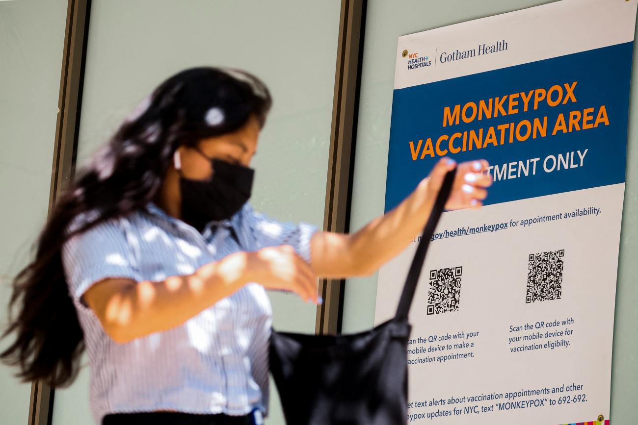 A woman with long dark hair in a light blue blouse and black mask searches for something in a black tote bag as she walks past a blue sign with orange letters reading "monkeypox vaccination area"