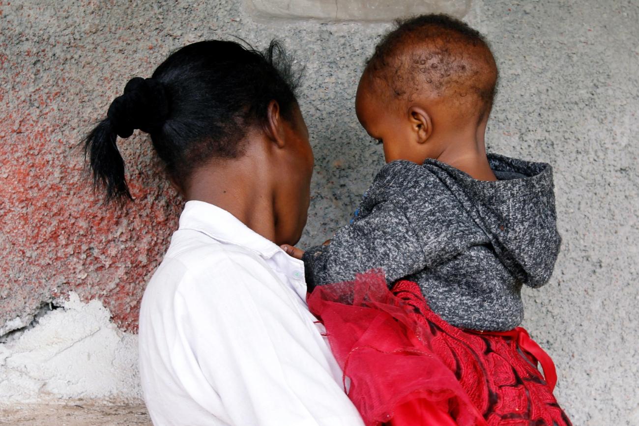 A young woman wearing a white shirt is photographed from behind holding her baby weraing a gray jumper