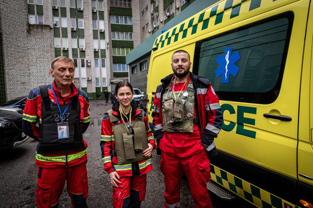 Health workers, two male and one female, wear red emergency outfits, stethoscopes, and bullet-proof vests as they  pose beside their yellow ambulance at a hospital in Kharkiv. 