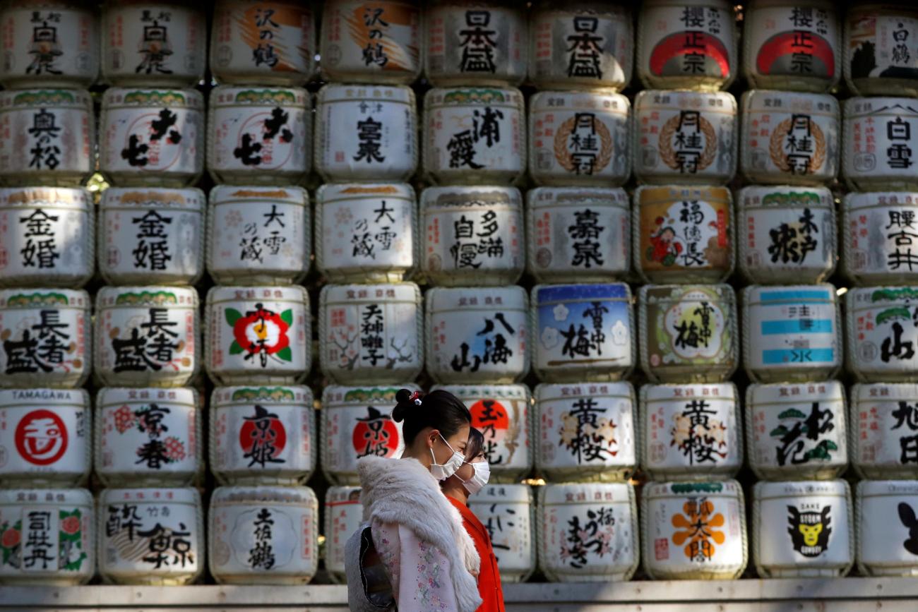 Kimono-clad women wearing protective face masks walk in front of white Japanese Sake barrels stacked in a grid decorated with colorful designs for the year-end and New-Year at Meiji Shrine