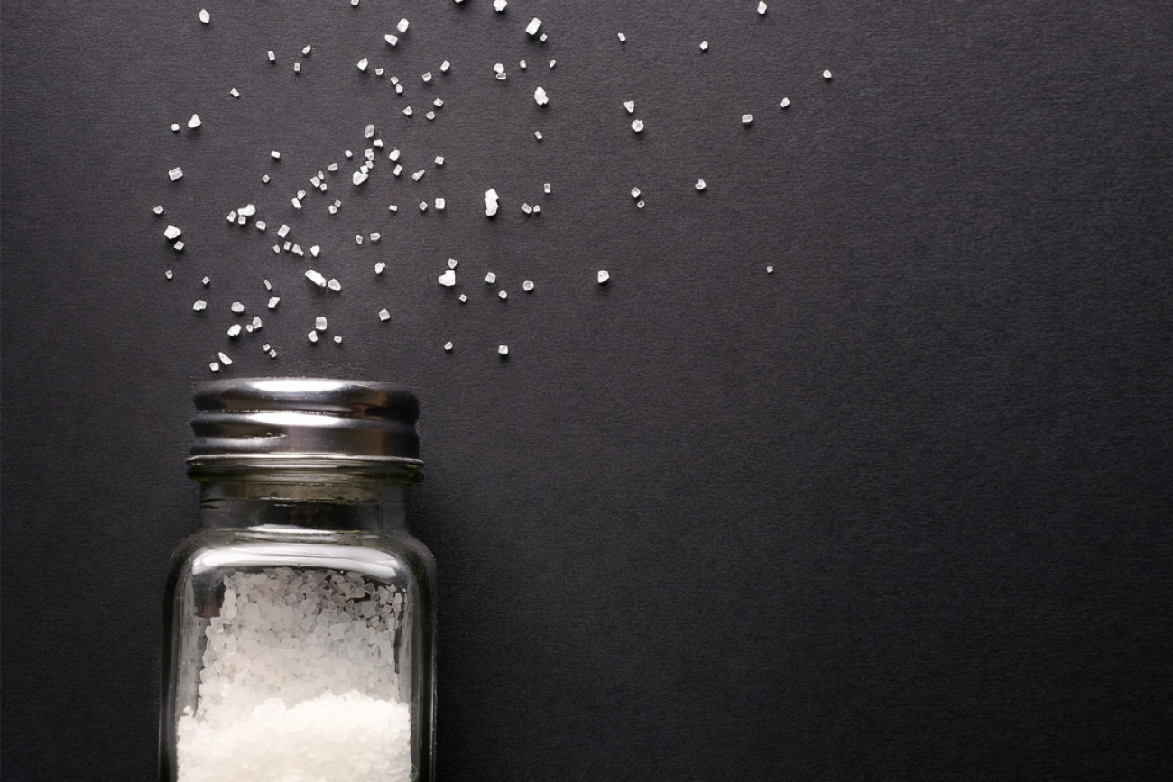 Natural, organic white sea salt pours from a fallen salt shaker onto a black table. 