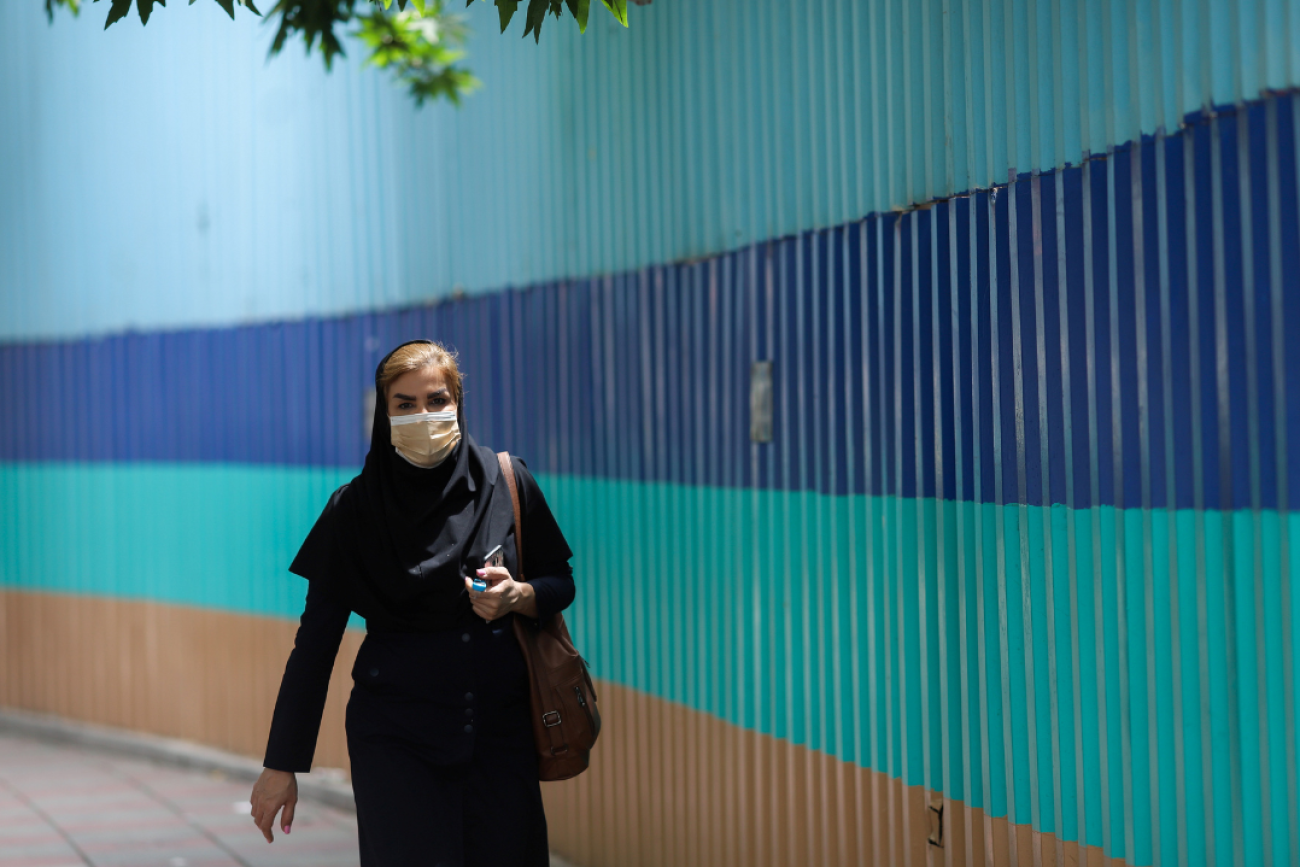 An woman walks along a street in Tehran, Iran, on May 26, 2021. Behind her stretches a wall with thick colorful horizontal stripes in shadess of blue and aqua.