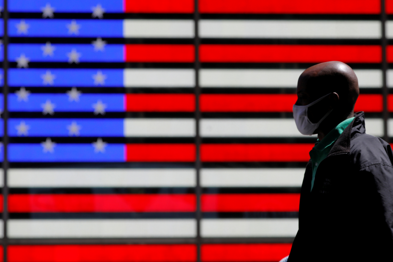 A giant red, white, and blue lit-up U.S. flag can be seen behind a person wearing a mask while walking in Times Square after the CDC announced new guidelines regarding outdoor mask wearing and vaccination during the COVID outbreak in New York City, New York, on April 27, 2021. 