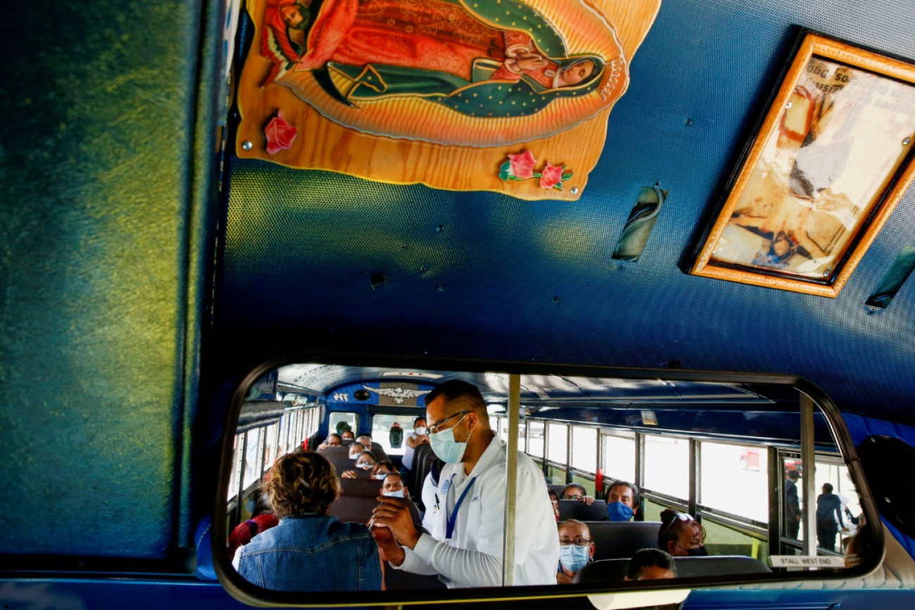 A rearview mirroron a bus reflects the image of assembly factory employees sitting in the bus waiting to receive COVID-19 vaccines. A health worker in a white medical coat is giving a vaccine to one of the women. Photo taken in Ciudad Juarez, Mexico, on May 24, 2021. 