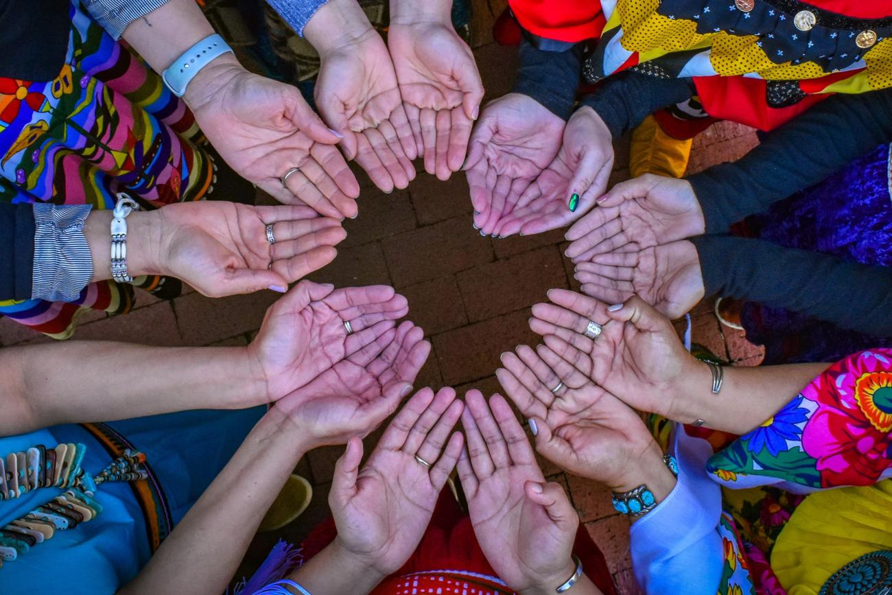 Changing Woman Initiative board and staff members, wearing traditional clothing from their respective tribes, connect with open hands to symbolize their acceptance of prayers and blessings.