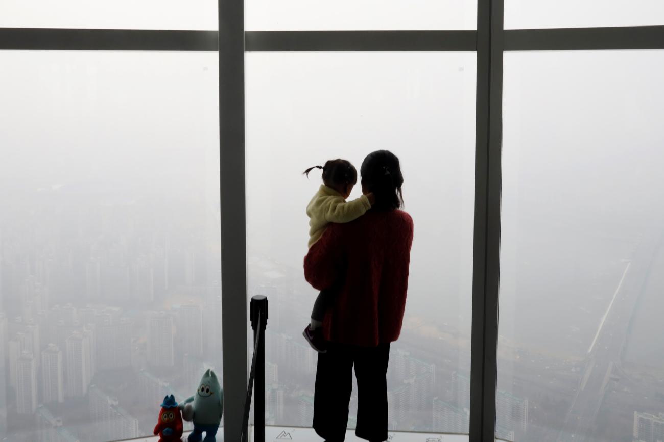 A woman holding her baby in her arms looks out a window at the city shrouded by a fine dust during a polluted day.