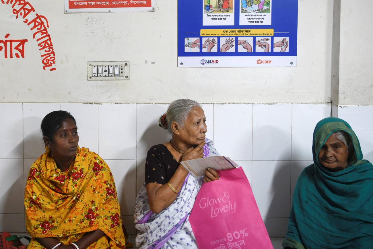 Patients wait for treatment as a USAID-funded health awareness poster hangs behind them, at the Hossainpur Upazila Health Complex, in Kishoreganj, Bangladesh.