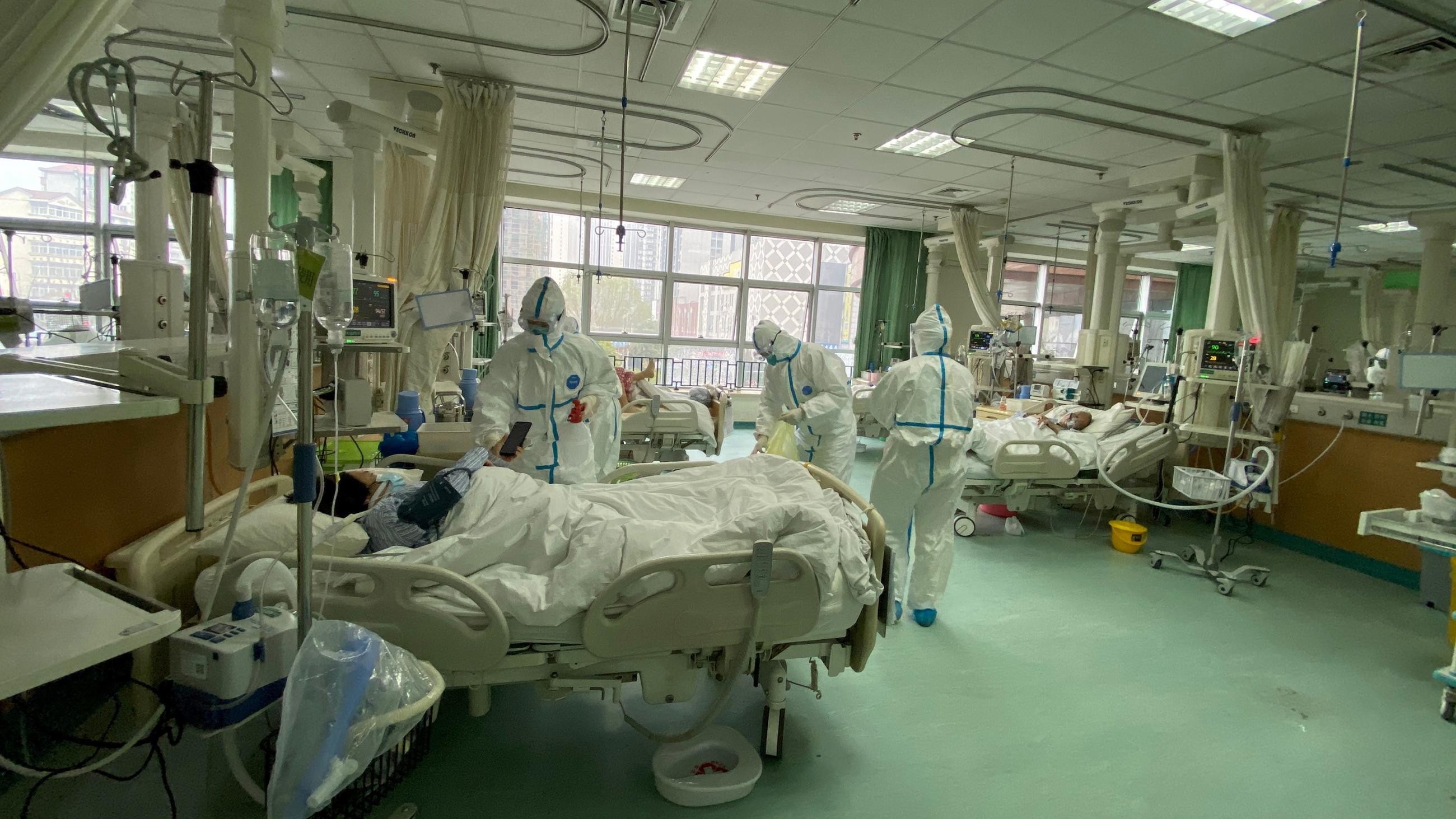 The picture shows a modern hospital ward with a patient in bed covered in protective gear as well as several health care workers covered head-to-toe in the same. 