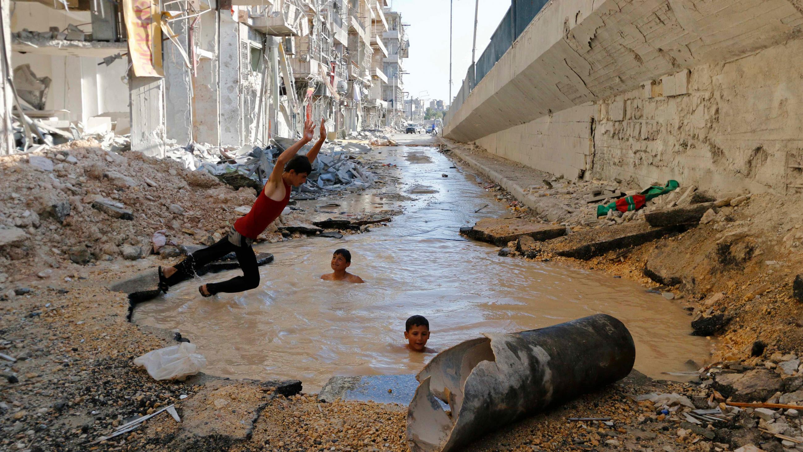 The photo shows a boy leaping in mid-air into a water-filled hole. The water is muddy brown, and two other boys can be seen swimming in the hole with their heads bobbing out. Surrounding them is rubble. This is a powerful image.