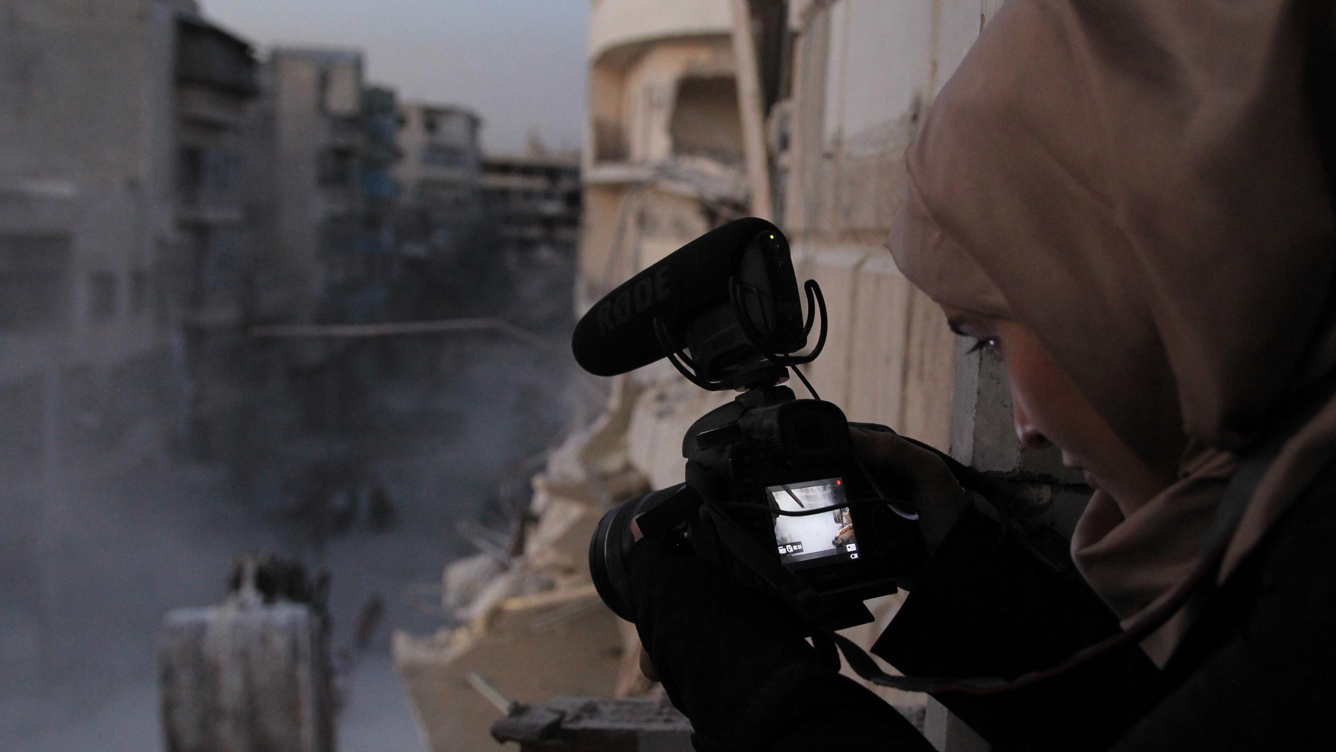 The picture shows the filmmaker from behind, as she captures a view of destruction from a high vantage overlooking the city street. 