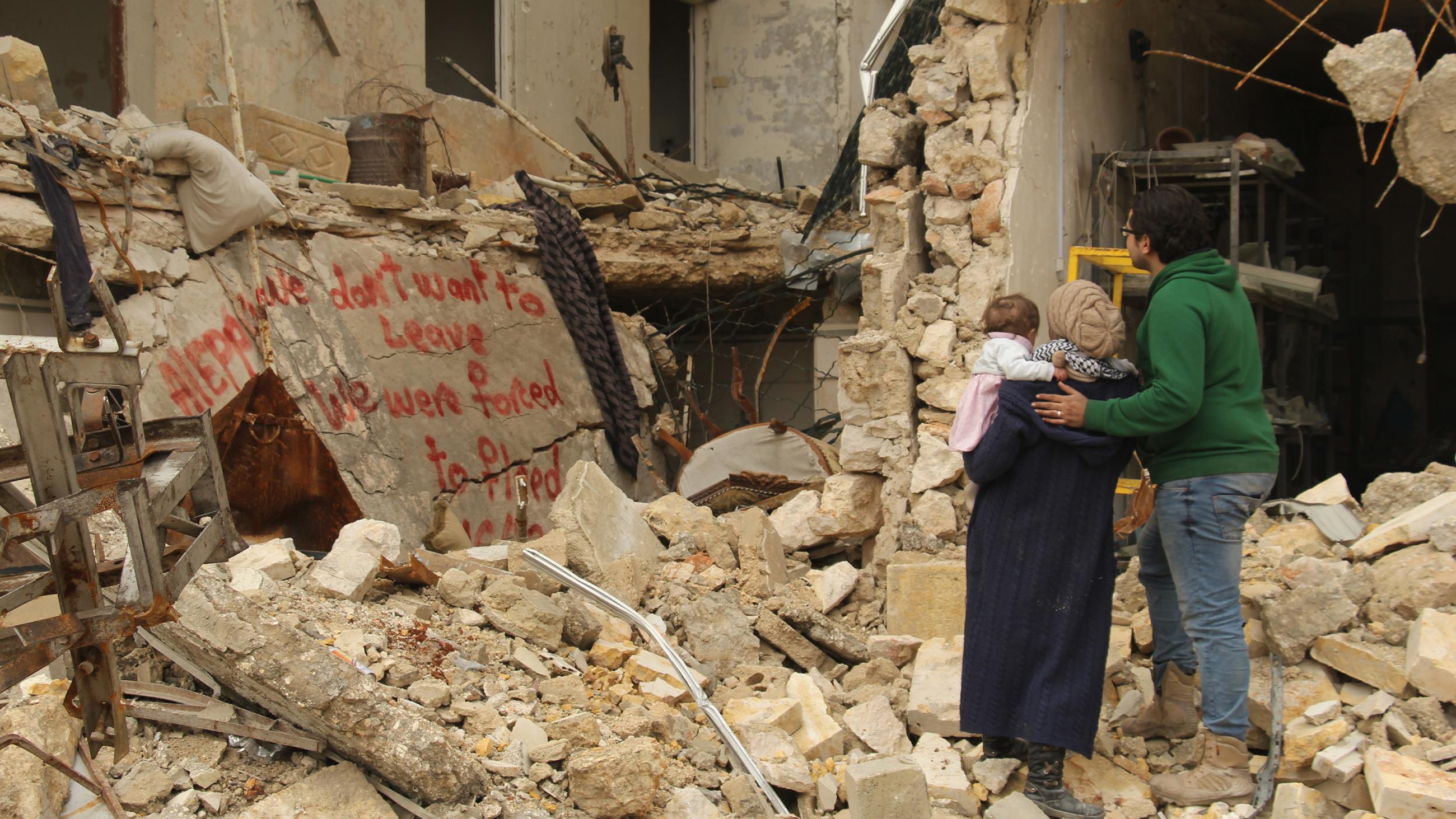 The picture shows the filmmaker holding her baby with her husband at her side. They are standing on top of rubble in front of a bombed-out building with twisted metal exposed and walls crumbled. Spray paint on a wall nearby reads, “We don’t want to leave / we were forced to flee.” 