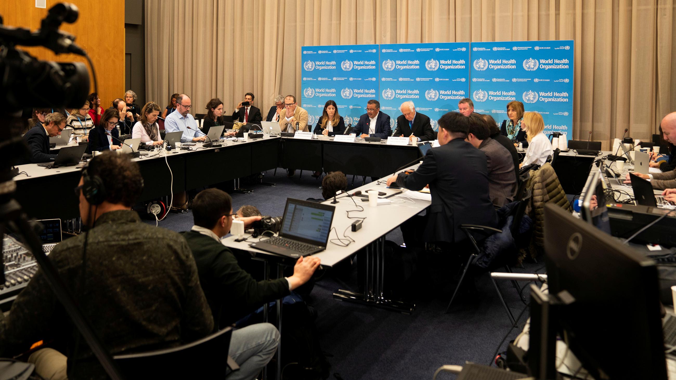 Picture shows a crowded press conference with officials seated at a long table and lots of reporters in the room.