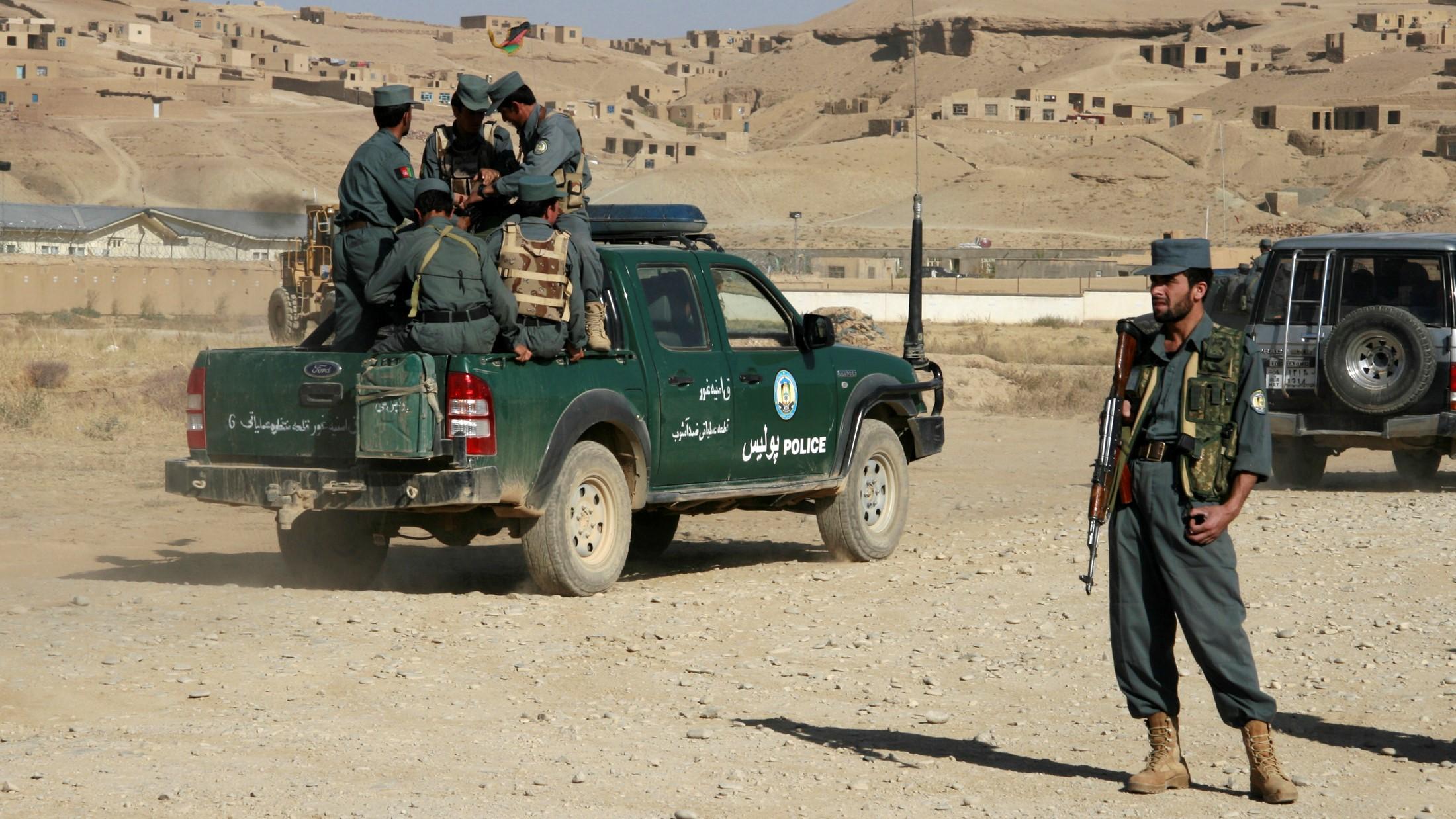 Photo shows a group of uniformed officers huddled in the back of a dark green pickup truck with "Police" written on the side. They are stationed in front of a village on a hill with several low-lying structures that appear to be built out of natural materials. One officer stands at attention away from the truck.
