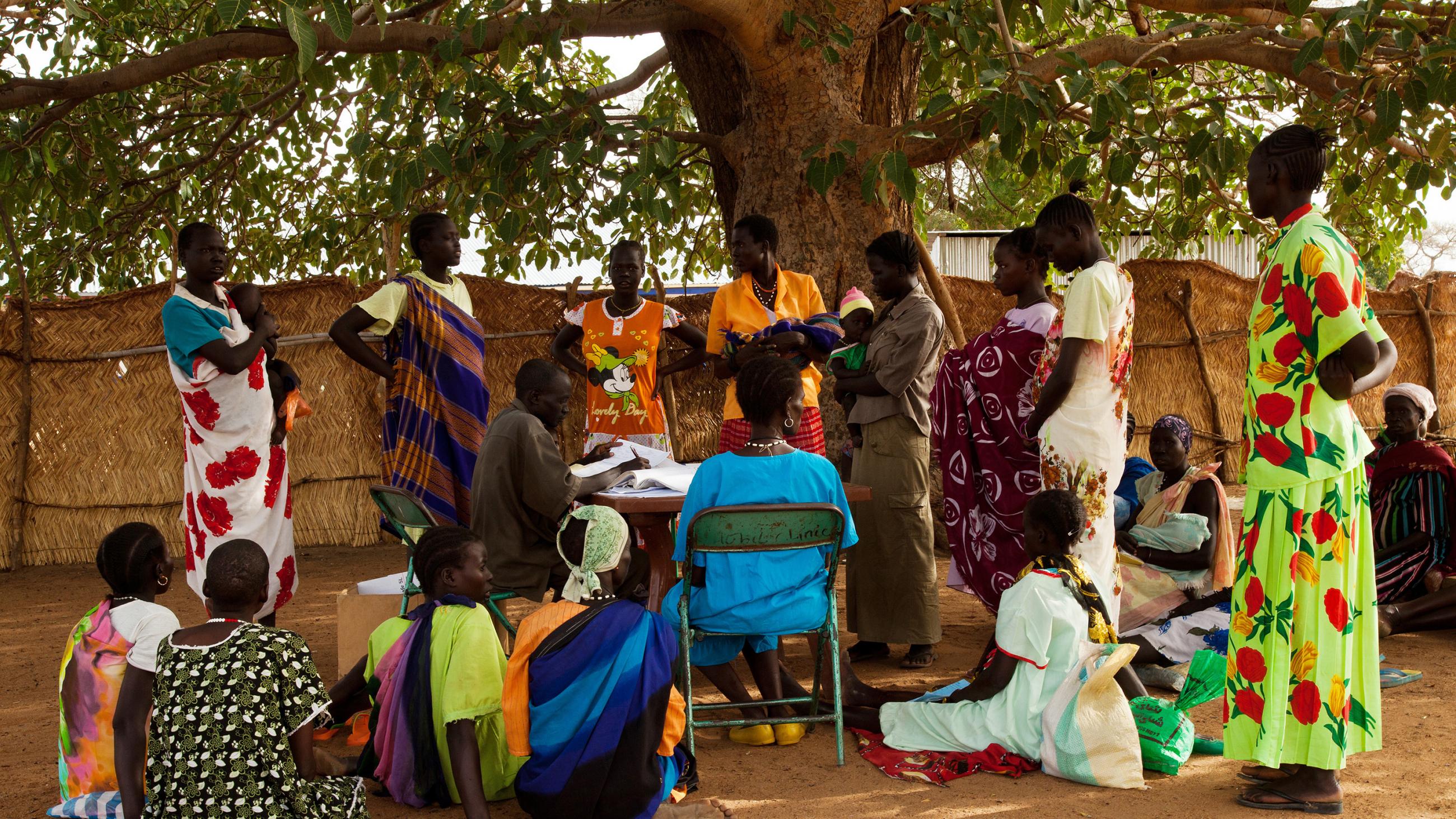 Picture shows a group of people, many of whom are wearing brightly coloured clothing, gathered under a tree, some sitting, some standing. 