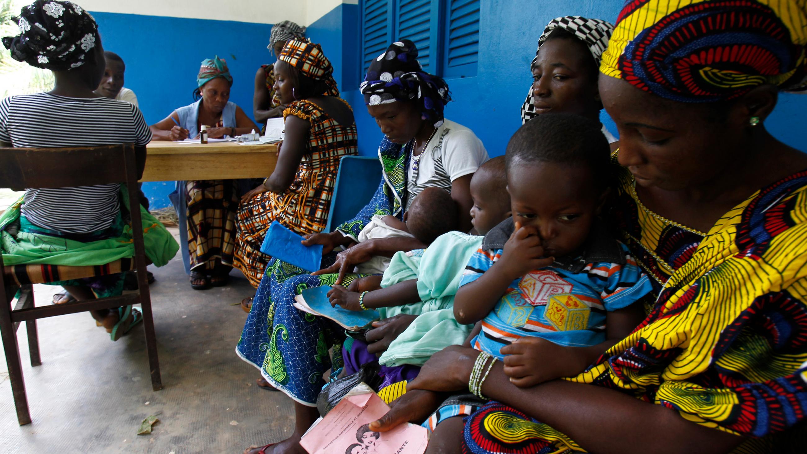  Image shows several women colorfully dressed holding young children and waiting on a bench. 