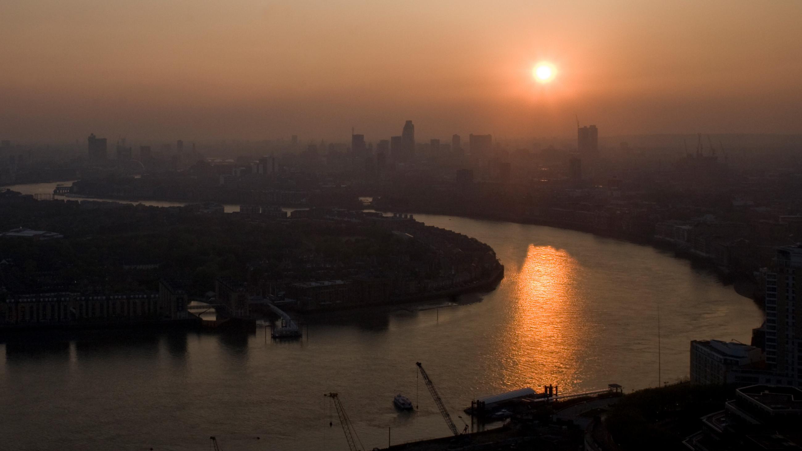The picture shows a high angle shot of London at dusk with the sun setting over the Thames. Smog obscures buildings in the distance. 