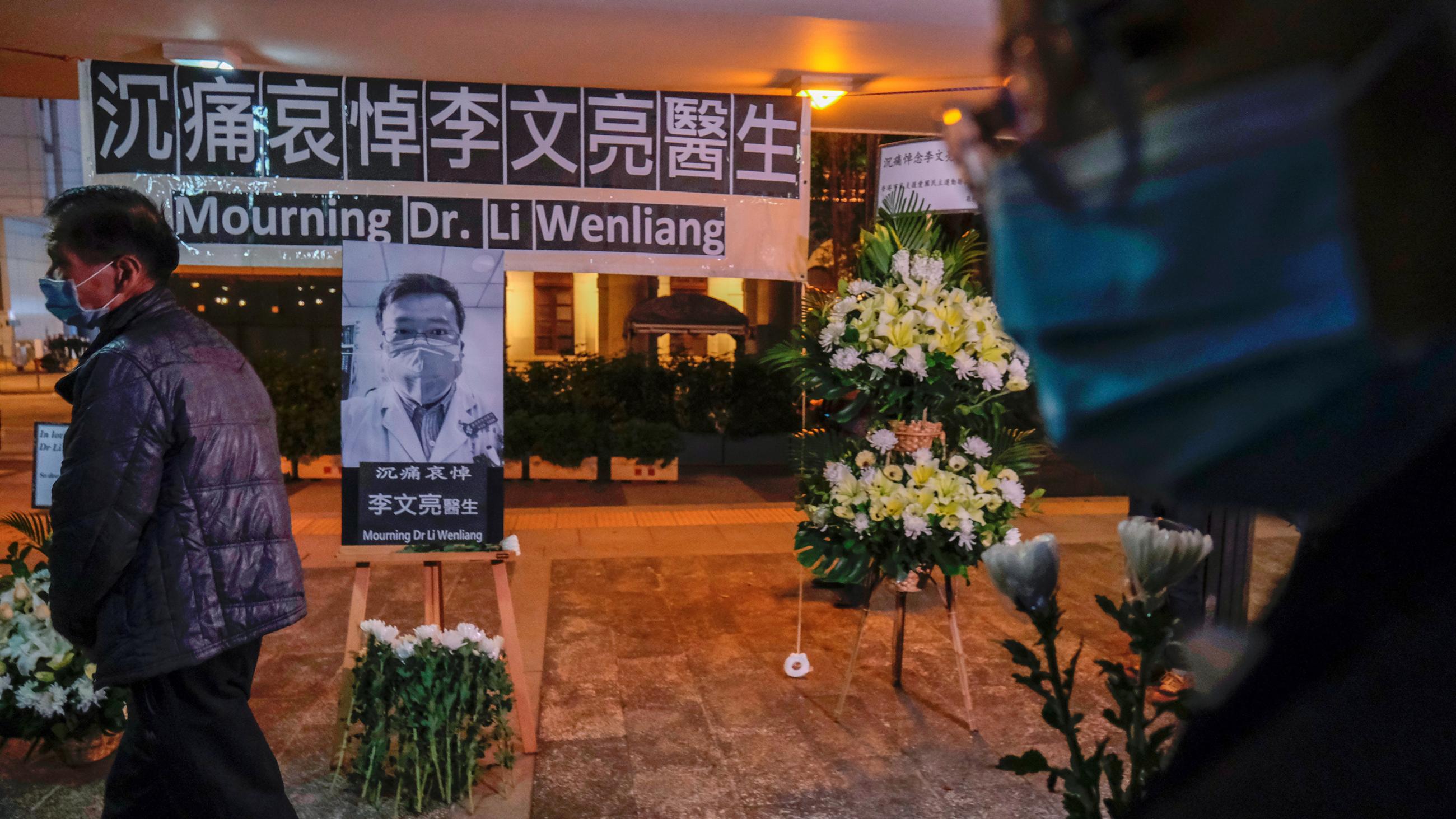 Image shows a few people wearing masks walking past a sort of shrine for the deceased doctor with lots of flowers, an enlarged photo, and some words written in Mandarin.