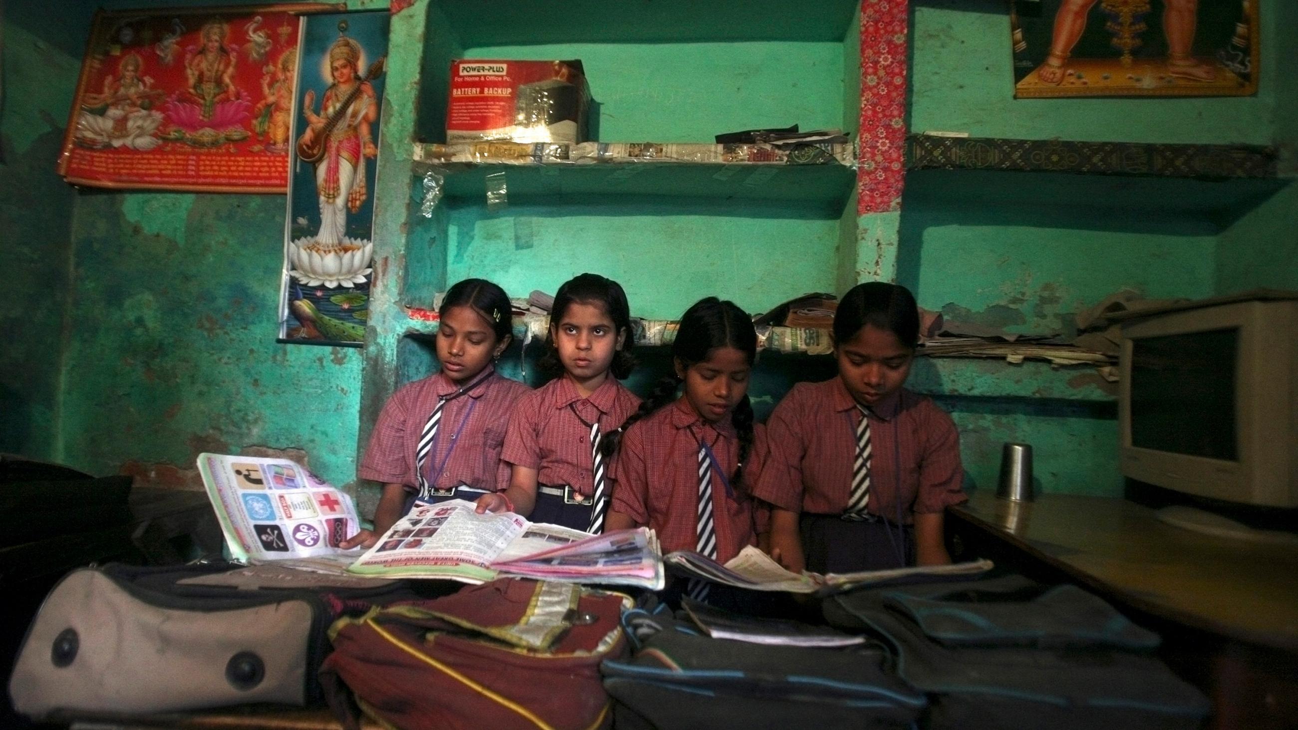 Image show four girls sitting close to each other with their heads buried in books, except one who looks up and off camera. The walls of the school are painted green. 