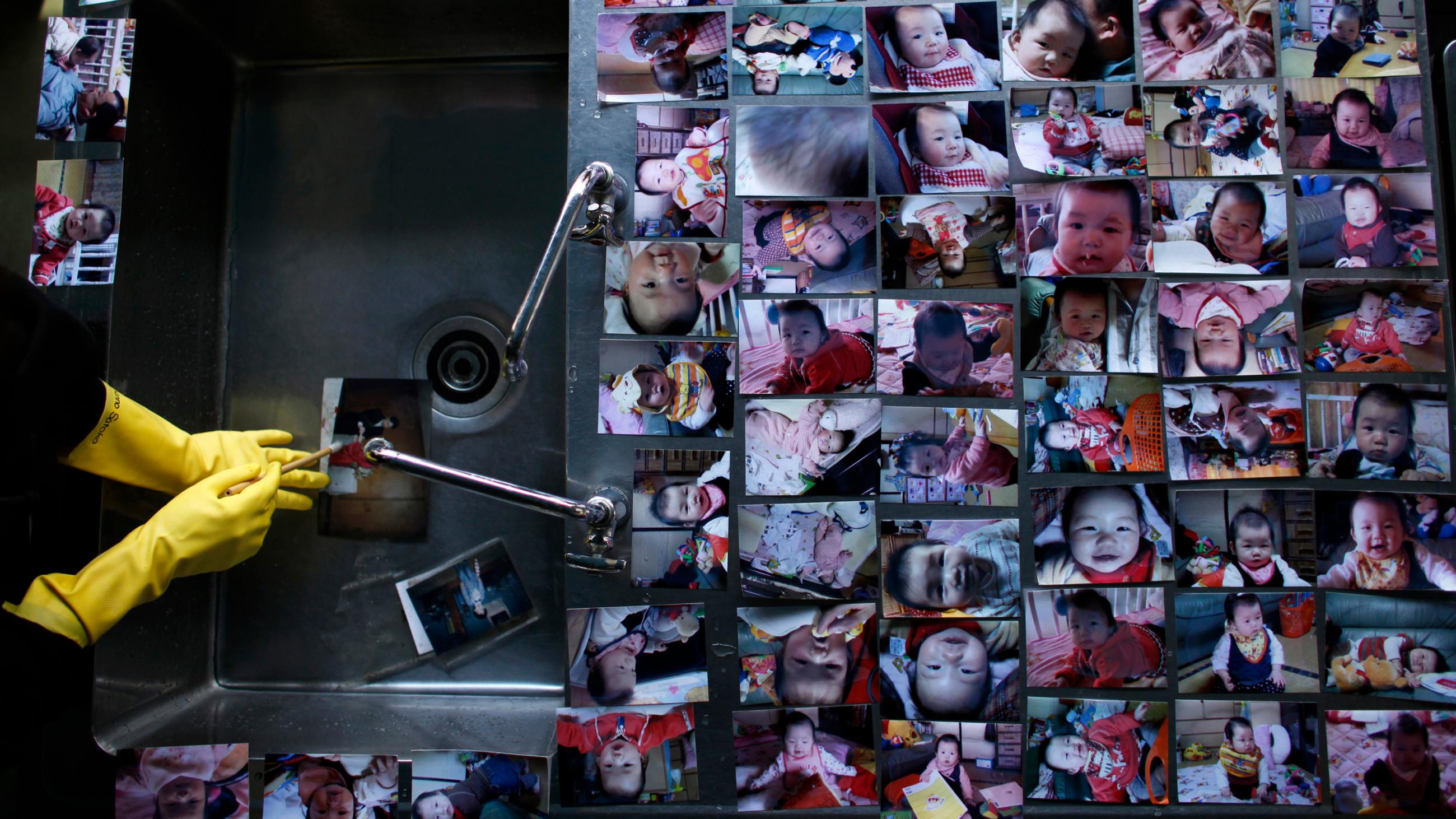 This is a stunning photo showing a metal sink with a broad metal shelf. A pair of hands wearing yellow dishwashing gloves is rinsing off a photo under the water. Meanwhile dozens of other photos, mainly of children, are laid out to dry on the edge of the sink. 