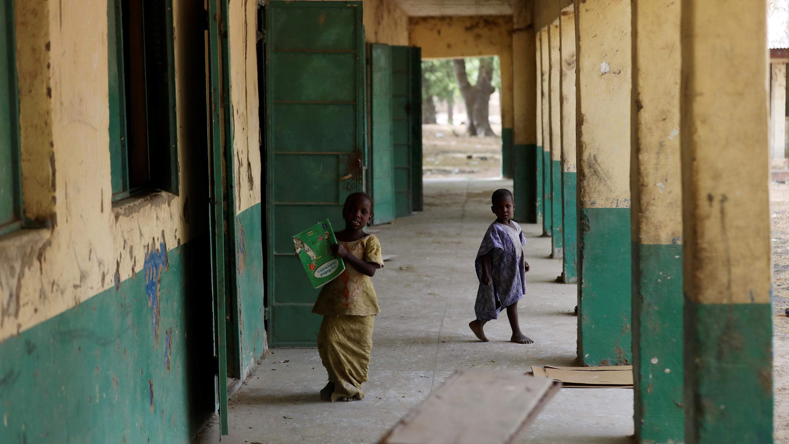  Picture shows two children walking along an outdoor corridor at a school with large cinder block columns painted green. 