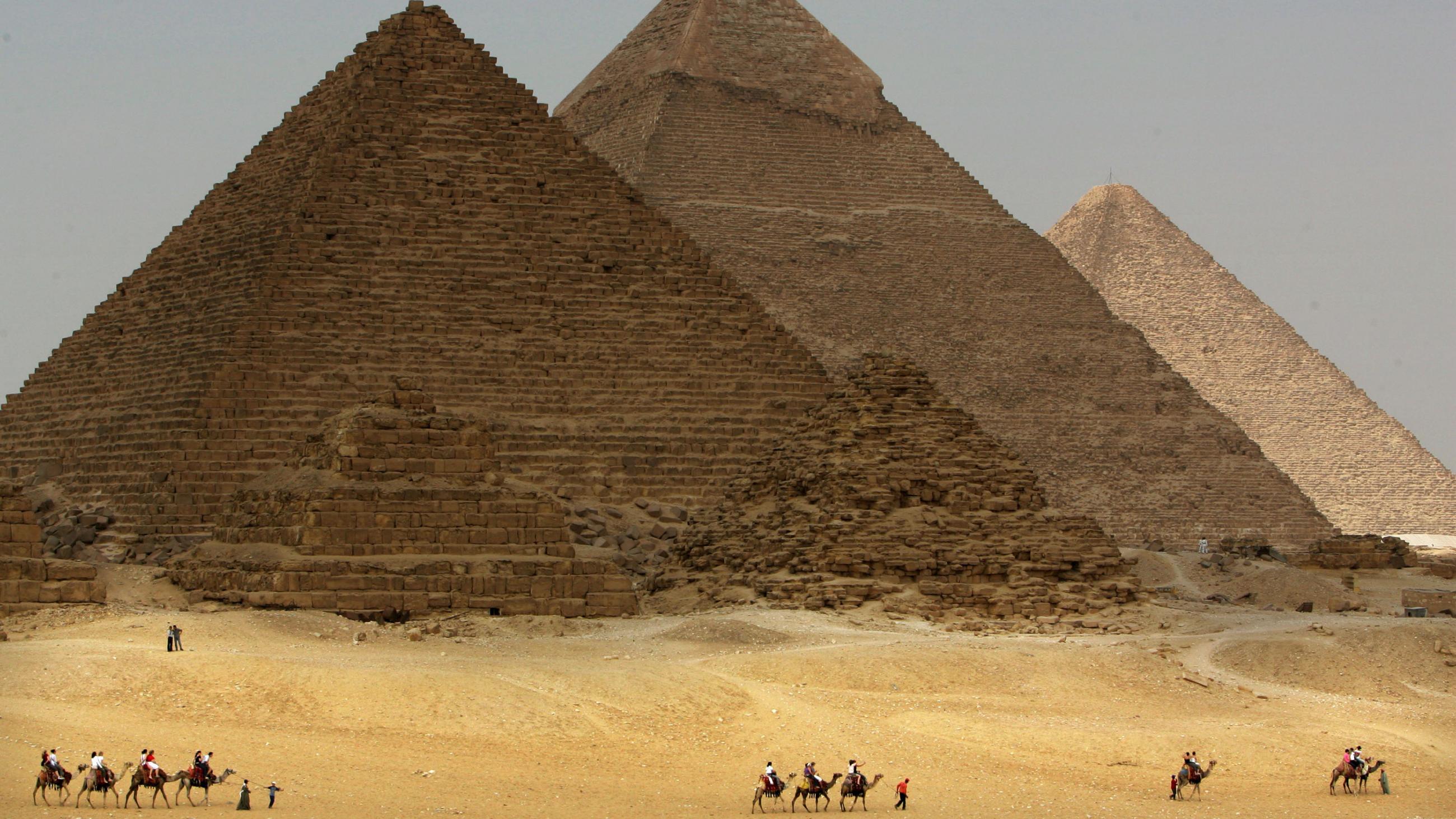 The picture shws the three pyramids off in the distance, taken from a three-quarters angle view. A long line of camels walks in front of the pyramids. 