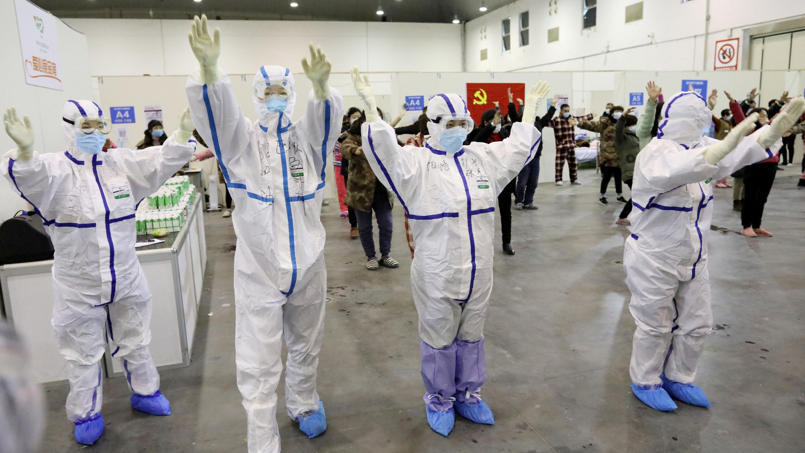 Picture shows a line of health care workers in full protective gear in front of other people who have masks, all with their arms in the air. 