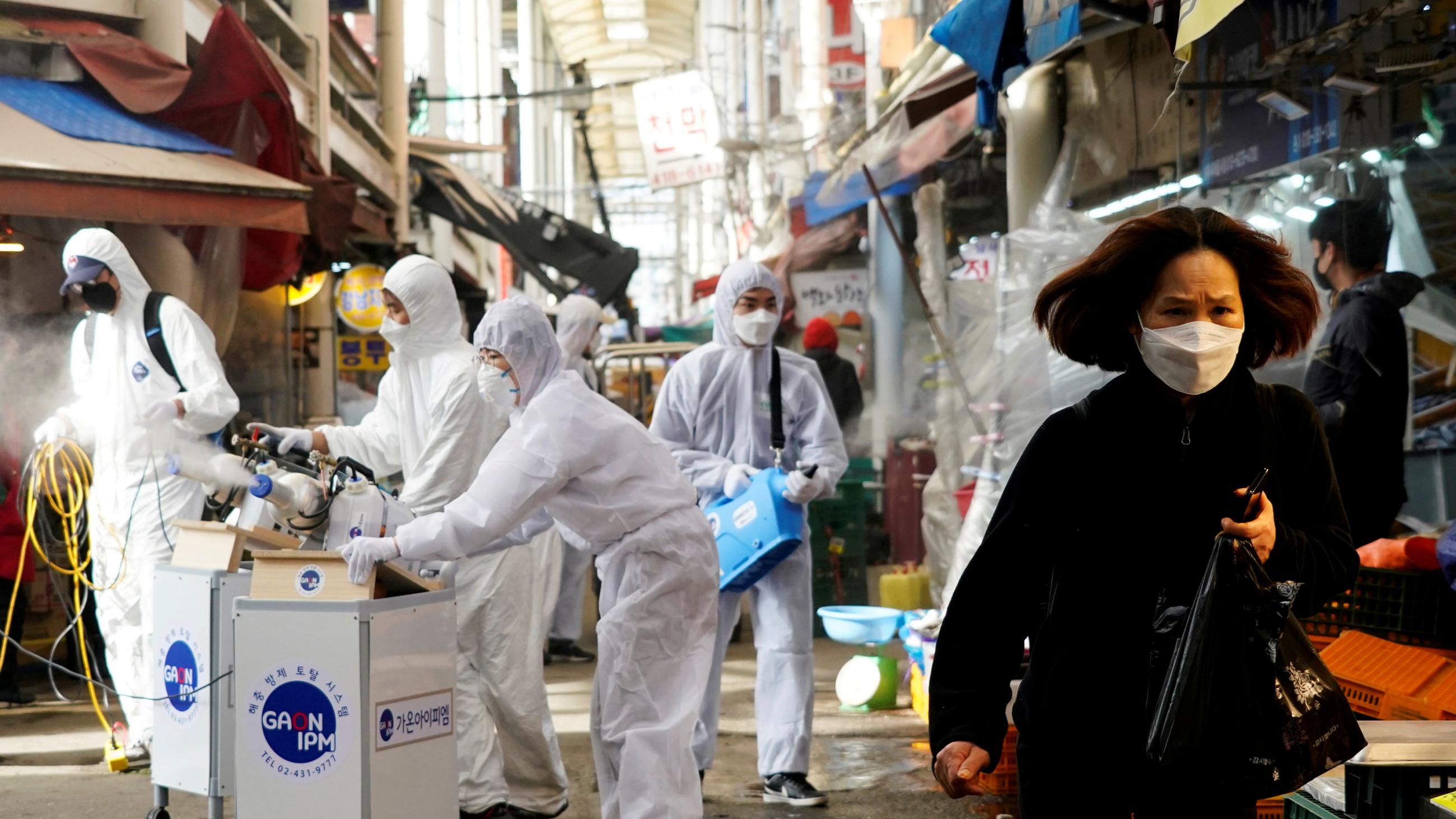 Image shows a woman walking through a crowded marketplace as a work crew teams up on an industrial sanitation machine. The look on her face suggests fear. 